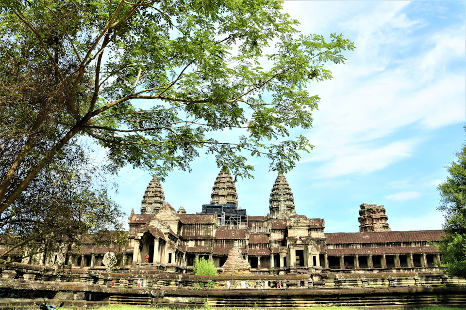 Ancient stone temple with tall spires, partially obscured by a leafy tree branch, under a bright blue sky.