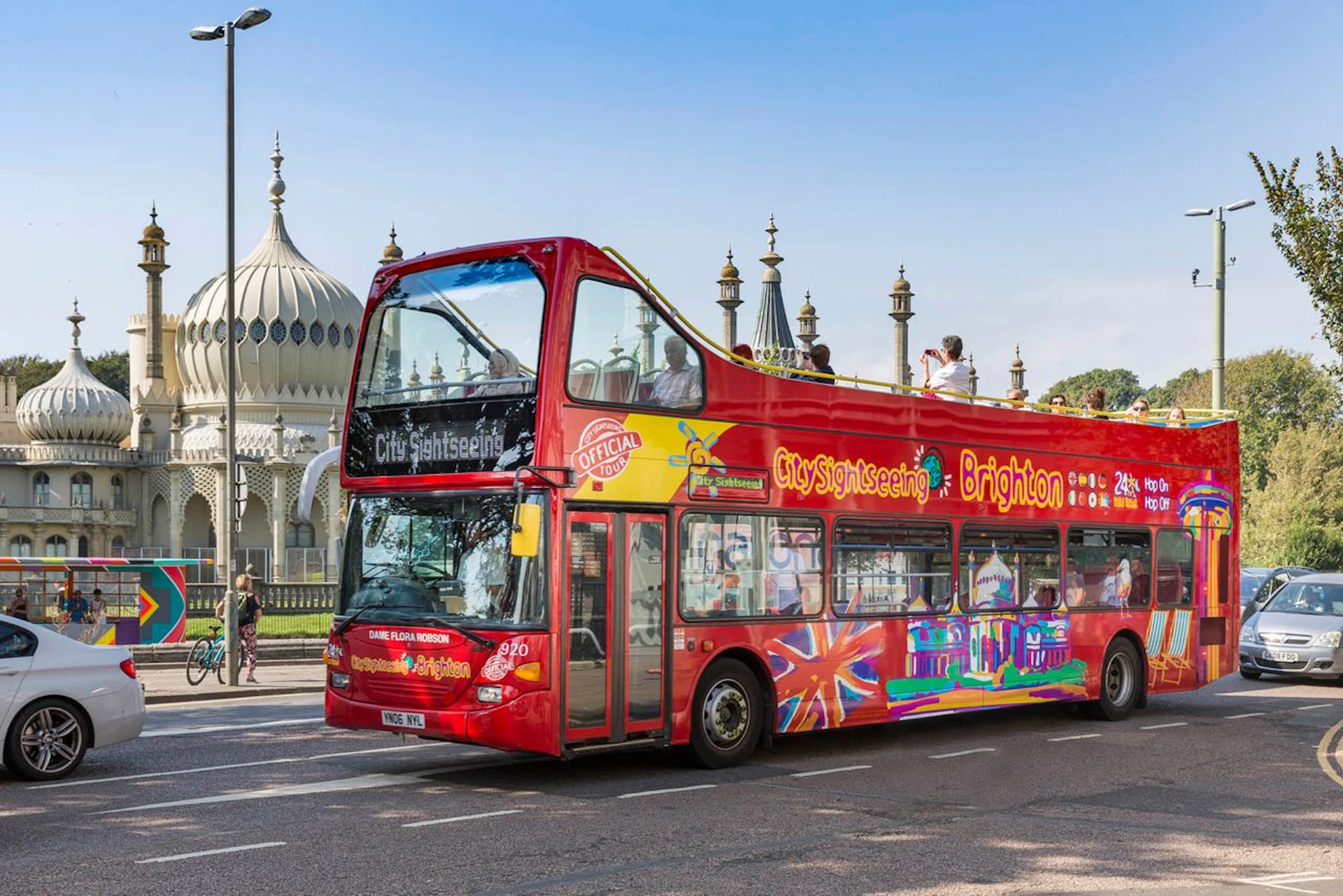 Red double-decker sightseeing bus in Brighton with tourists on the top deck, passing by a building with domed architecture.