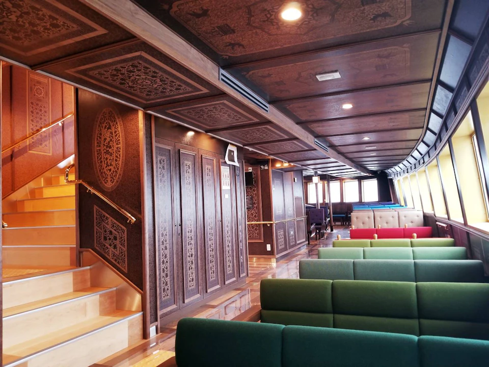 An elegant interior of a ship or building, featuring wooden paneling with intricate carvings, green and red seating, and stairs.