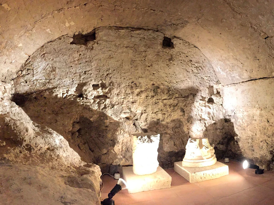 A dimly lit room with rough stone walls, featuring two illuminated stone artifacts on pedestals and spotlights on the floor.