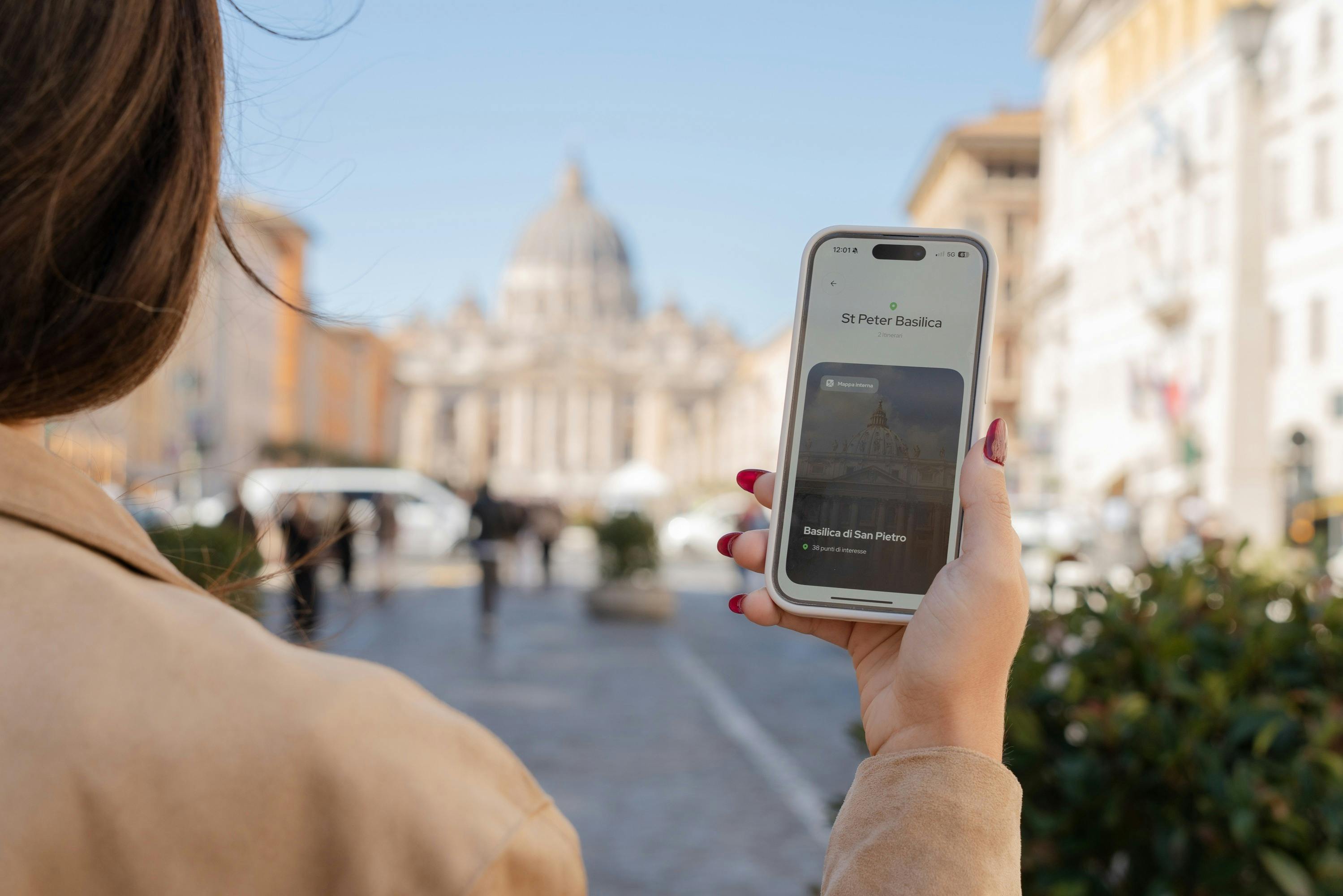 A hand holding a phone showing "St. Peter Basilica" with the actual basilica visible blurred in the background.