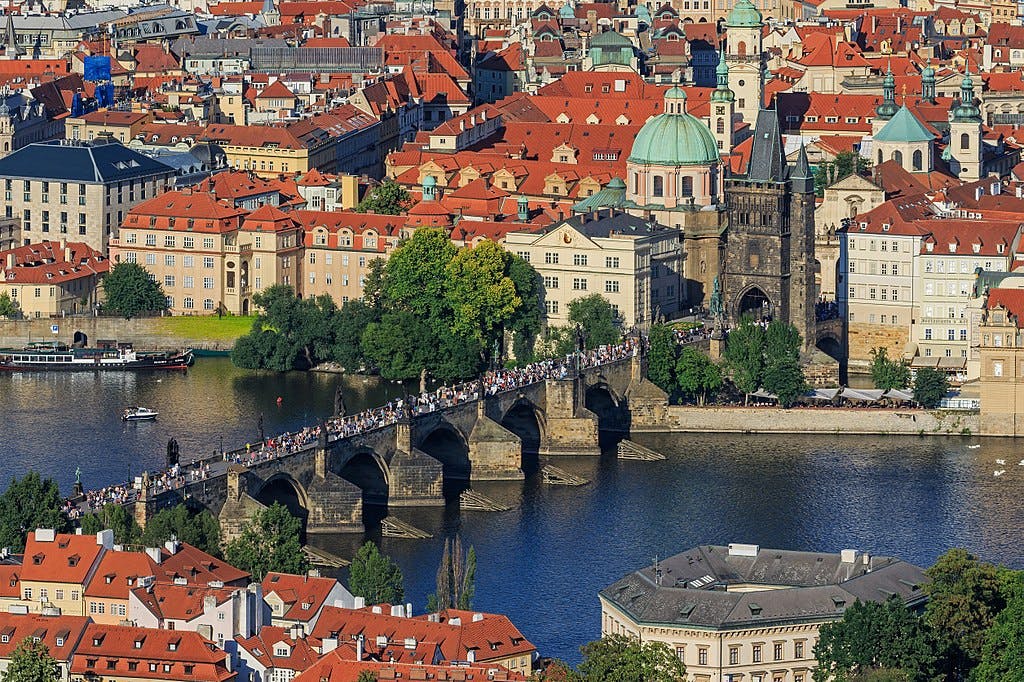Historic stone bridge over a river filled with pedestrians. Cityscape with buildings, red rooftops, and a domed church in the background.