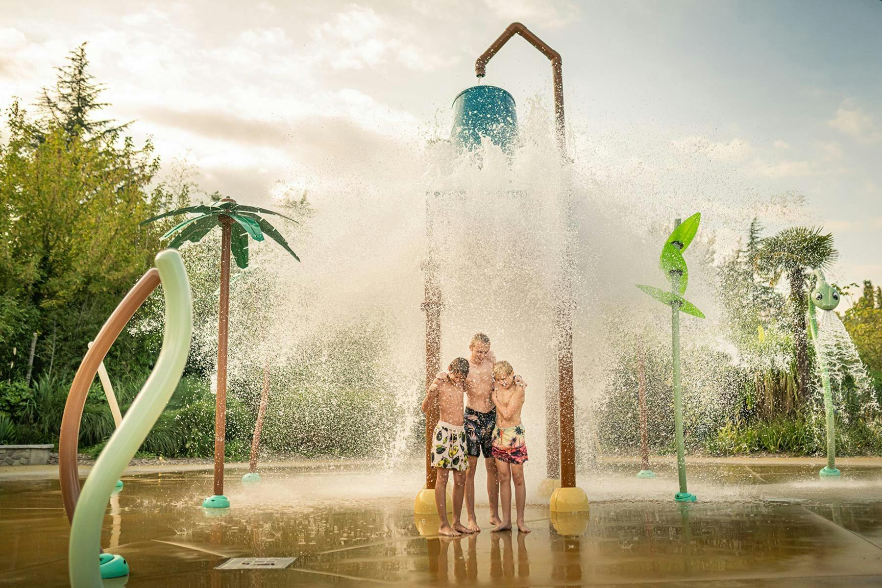 Three children stand under a large splash bucket dumping water at a water play area with tropical-themed decorations.