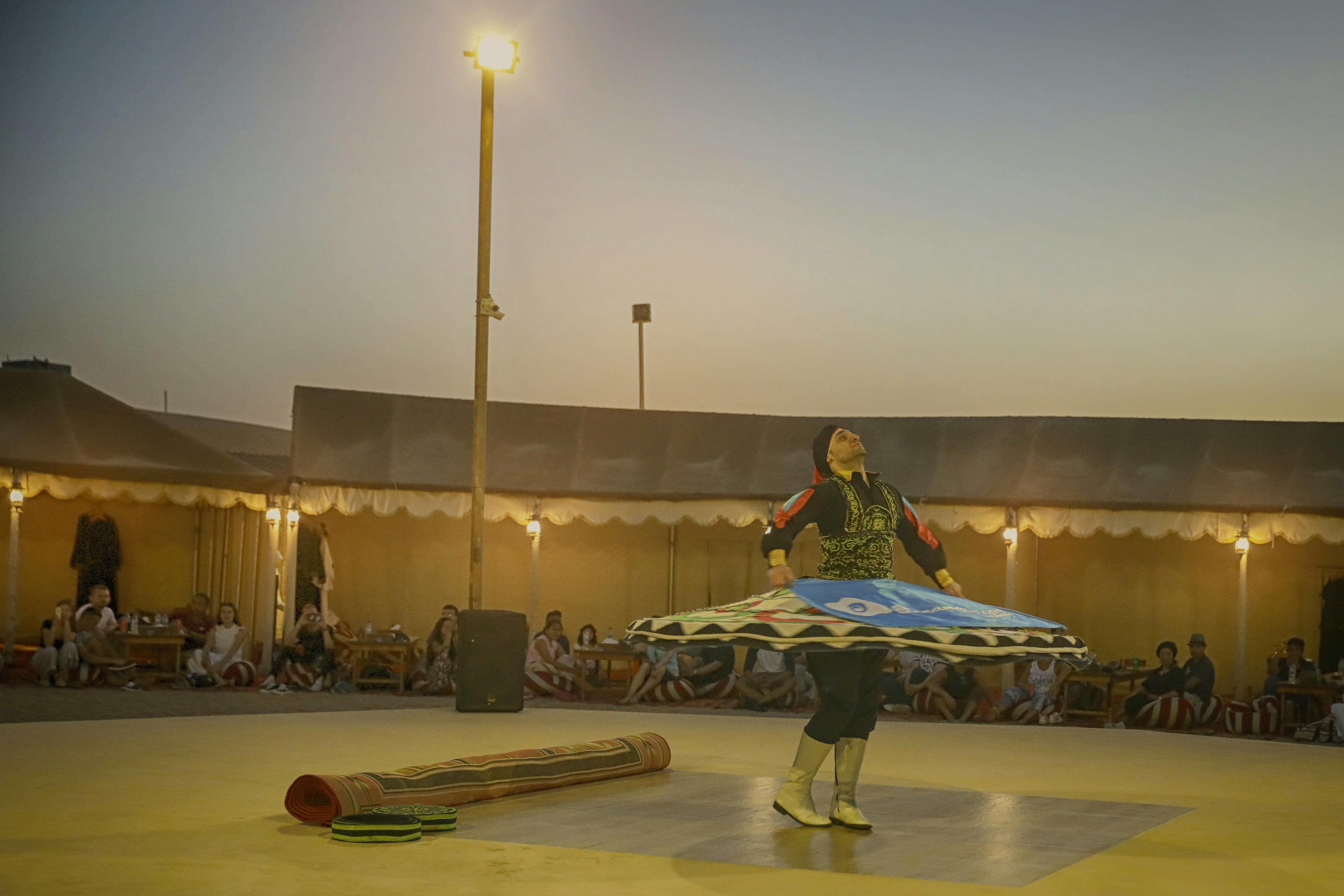 Umělec se otáčí v tradičním oděvu pod večerní oblohou, obklopen diváky pod širým nebem.