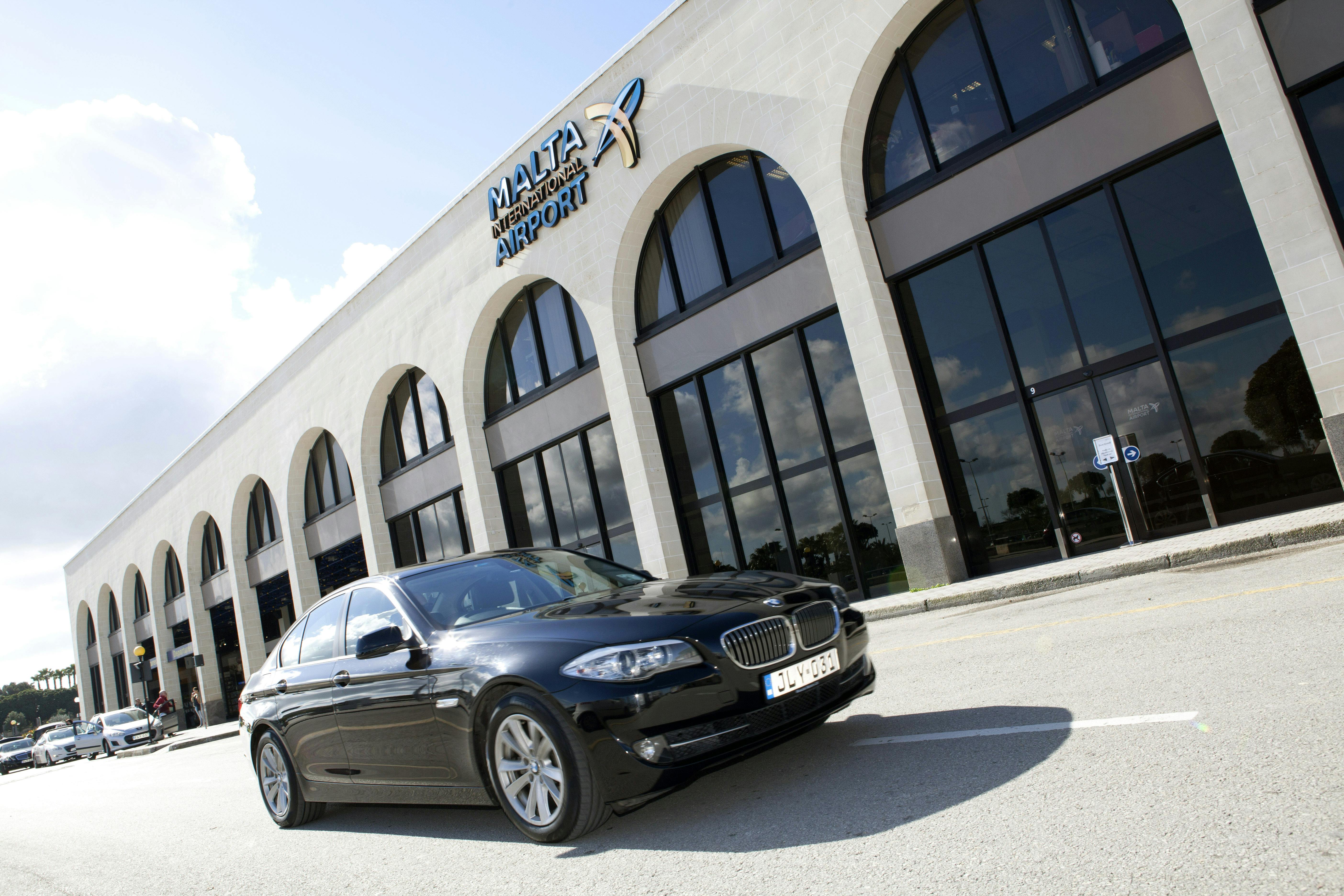 Černé auto zaparkované před budovou s klenutými okny a nápisem „Malta International Airport“.
