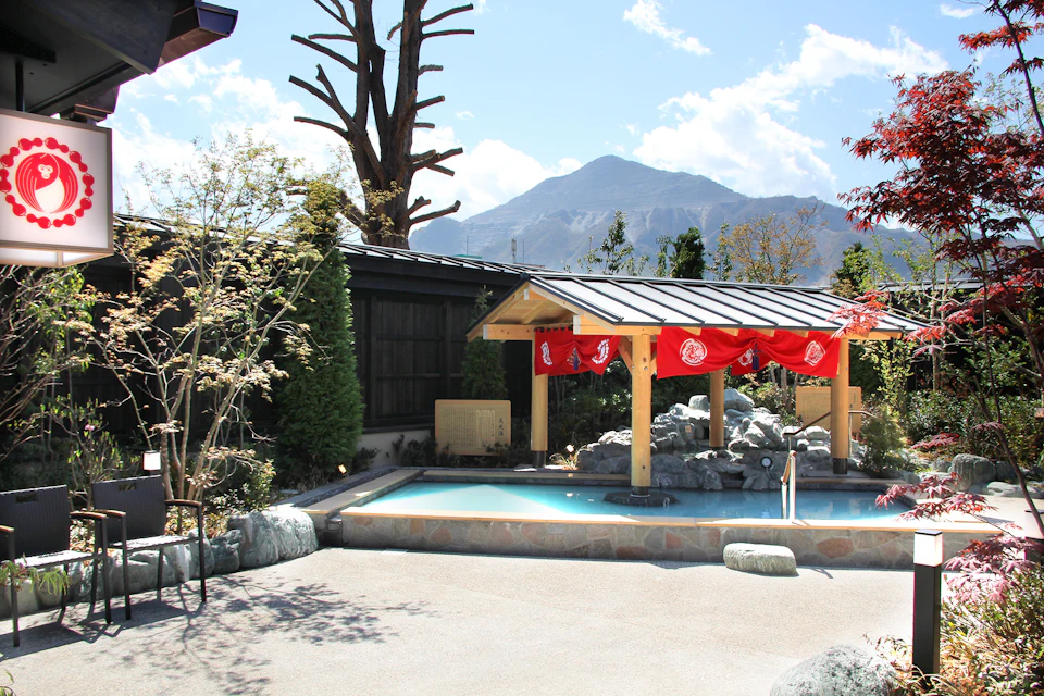 Outdoor hot spring bath with a wooden shelter and red banners, surrounded by trees and mountains in the background.