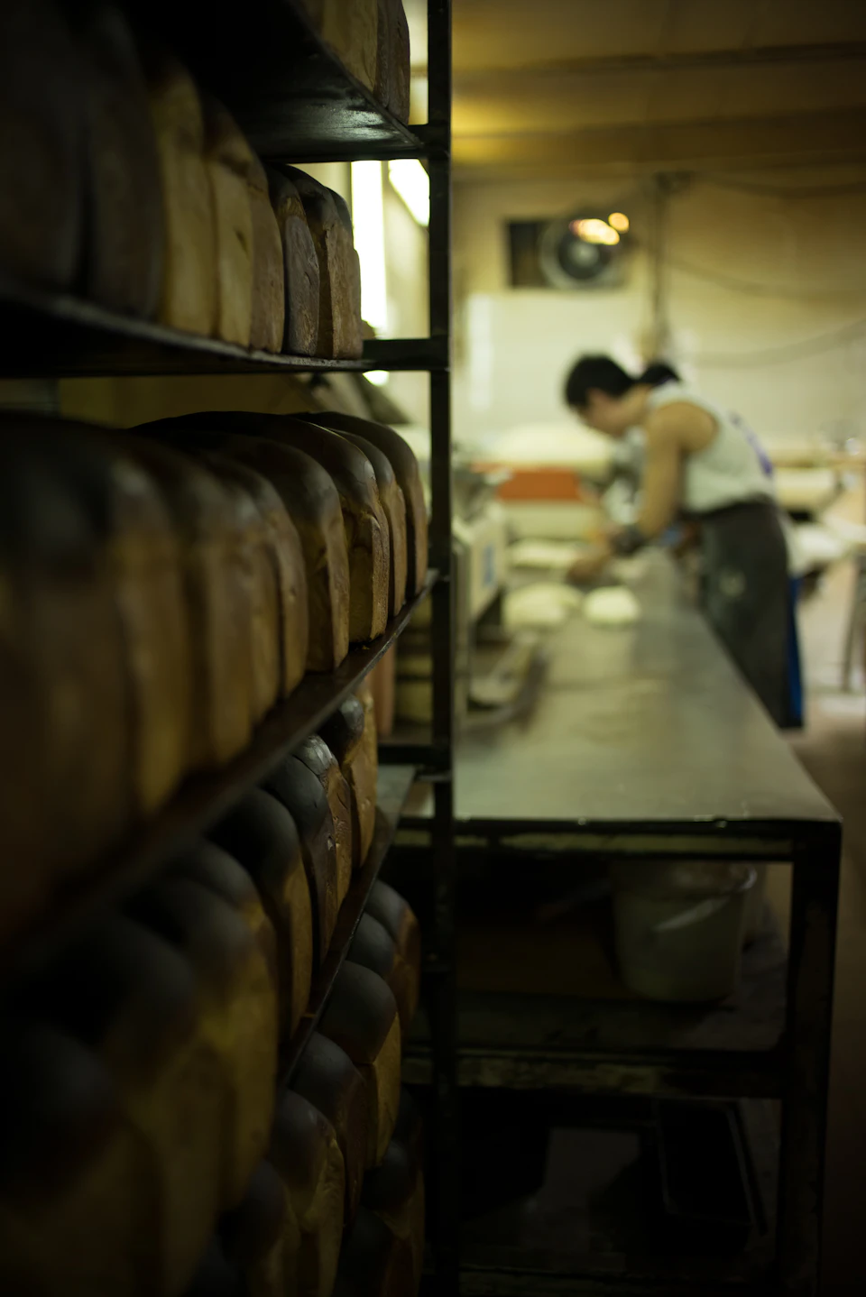 Loaves of bread on shelves in the foreground, with a baker working in the background at a long table in a dimly lit bakery.