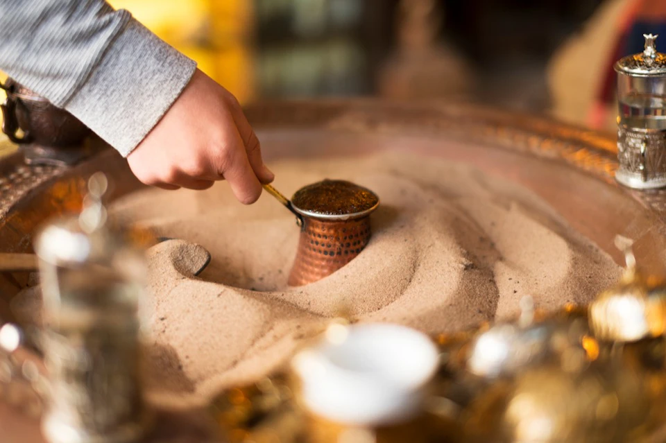 A hand holding a metal tool partially submerged in a pot filled with sand, with a blurred background.