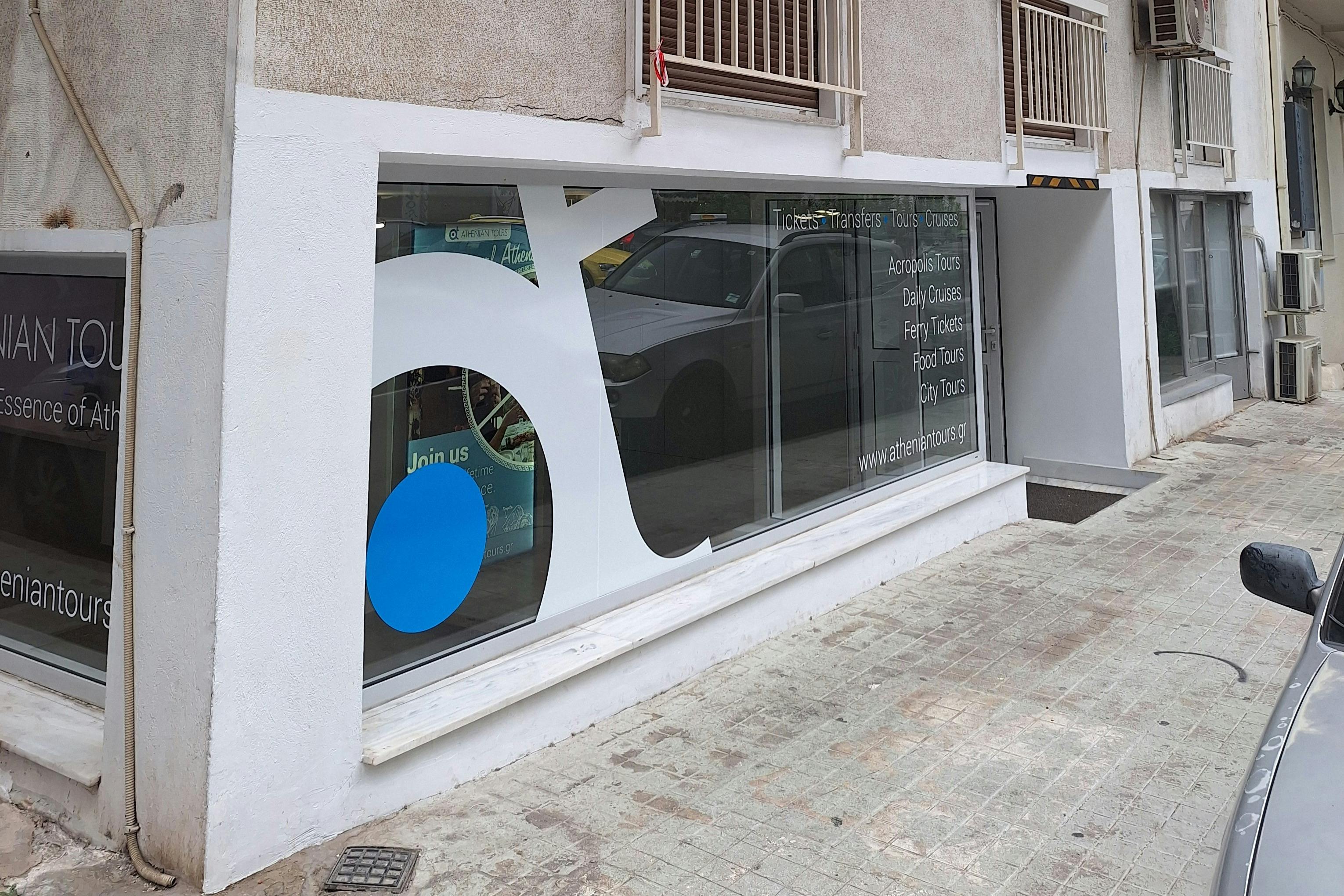 Γυάλινη βιτρίνα με μεγάλο λευκό και μπλε λογότυπο, με κείμενο που διαφημίζει διάφορες περιηγήσεις και υπηρεσίες, με αντανάκλαση αυτοκινήτου.