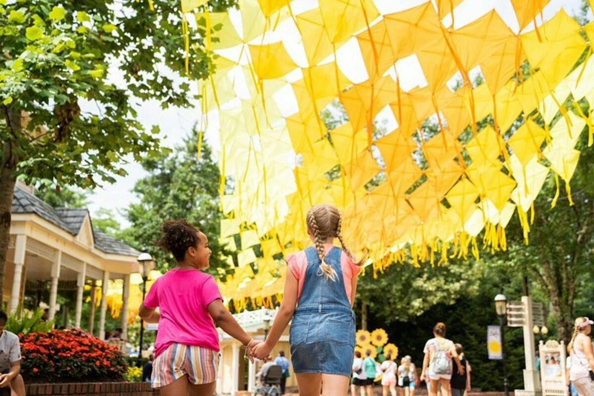 Two children walk hand in hand under a canopy of yellow decorations, surrounded by greenery and flowers in a park.