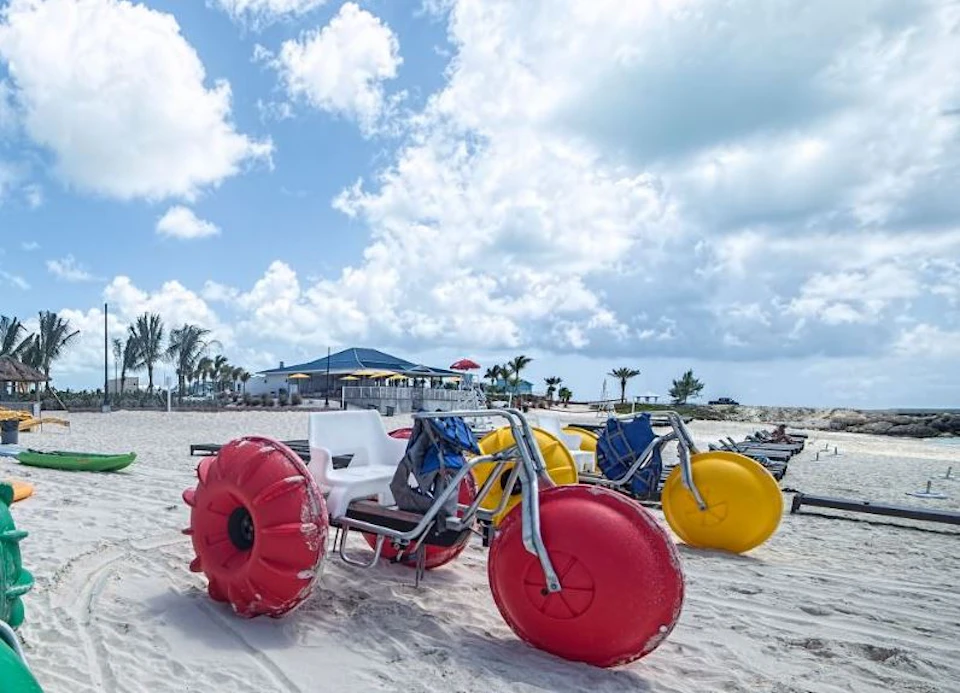 Beach scene with oversized tricycles featuring large red and yellow wheels on the sand, under a partly cloudy sky.