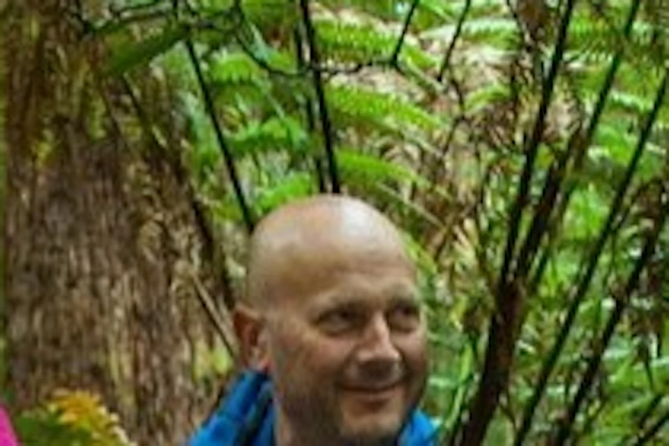 Three people in outdoor clothing standing in a lush forest, one pointing ahead while others listen. Logs and greenery surround them.