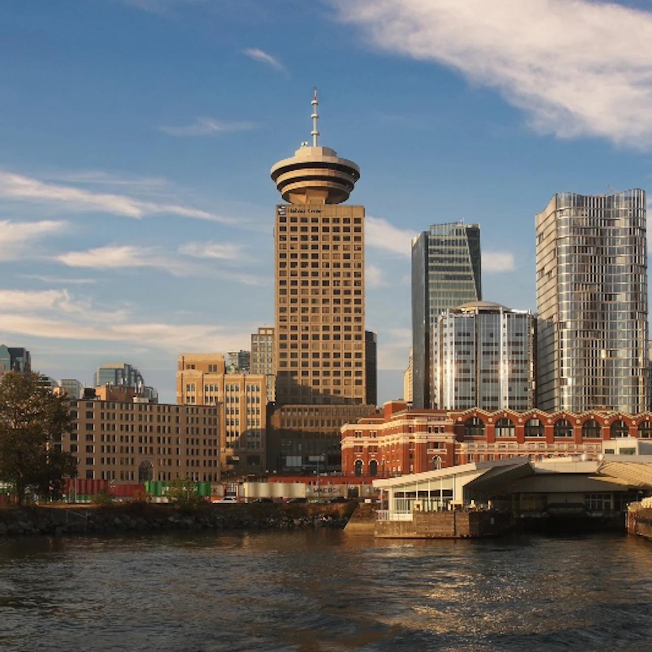 City skyline with a prominent round-topped tower and various skyscrapers, visible waterfront and bridge under a partly cloudy sky.