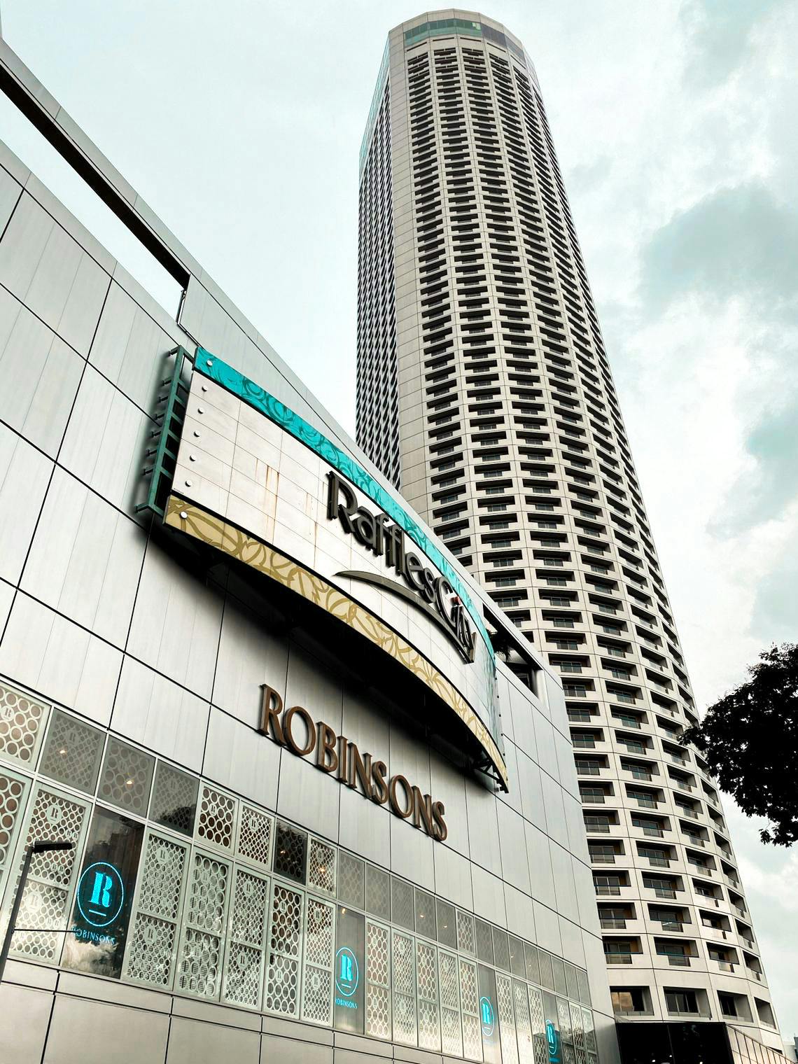 Alto grattacielo dietro la facciata di un edificio moderno con le insegne di "Raffles City" e "Robinsons". Rami di alberi sulla destra.