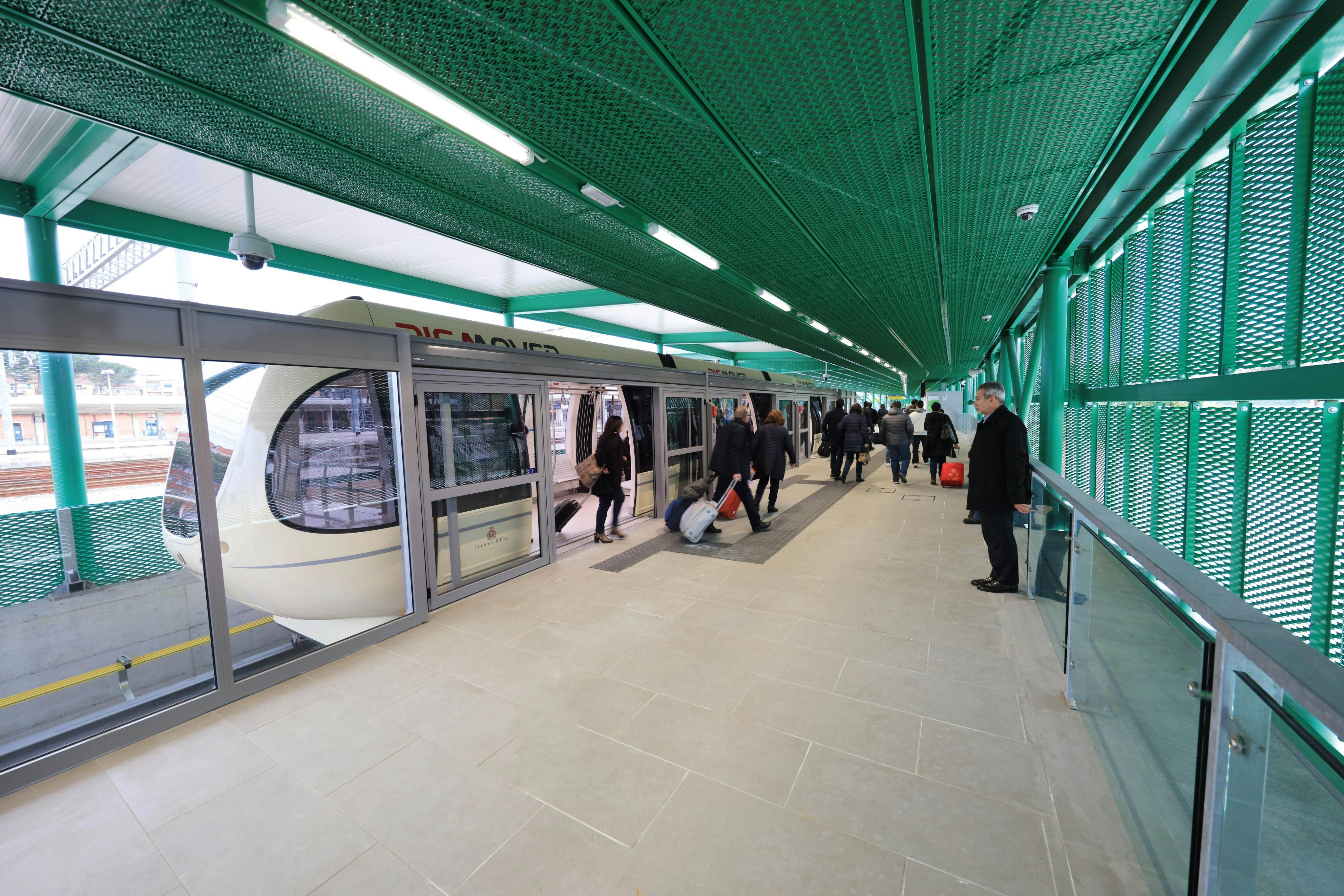 乘客在带有绿色结构元素的室内车站上下白色电车。有些人携带行李。