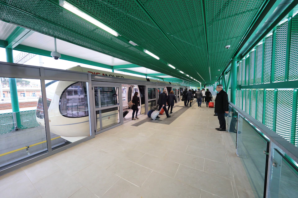 Passengers boarding and exiting a white tram at an indoor station with green structural elements. Some carry luggage.
