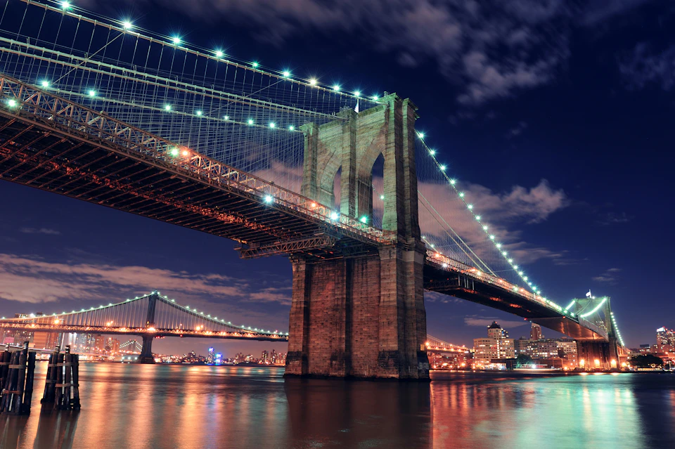 Two illuminated suspension bridges over a river against a city skyline at night, with reflections on the water and a partly cloudy sky.