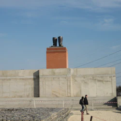 Två personer som går nära en stor betong- och tegelstruktur toppad med stora statyer av stövlar, under en klarblå himmel.