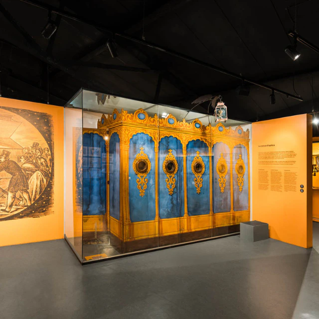 An ornate blue and gold carousel organ display in a museum with orange walls and spotlights illuminating the exhibit.