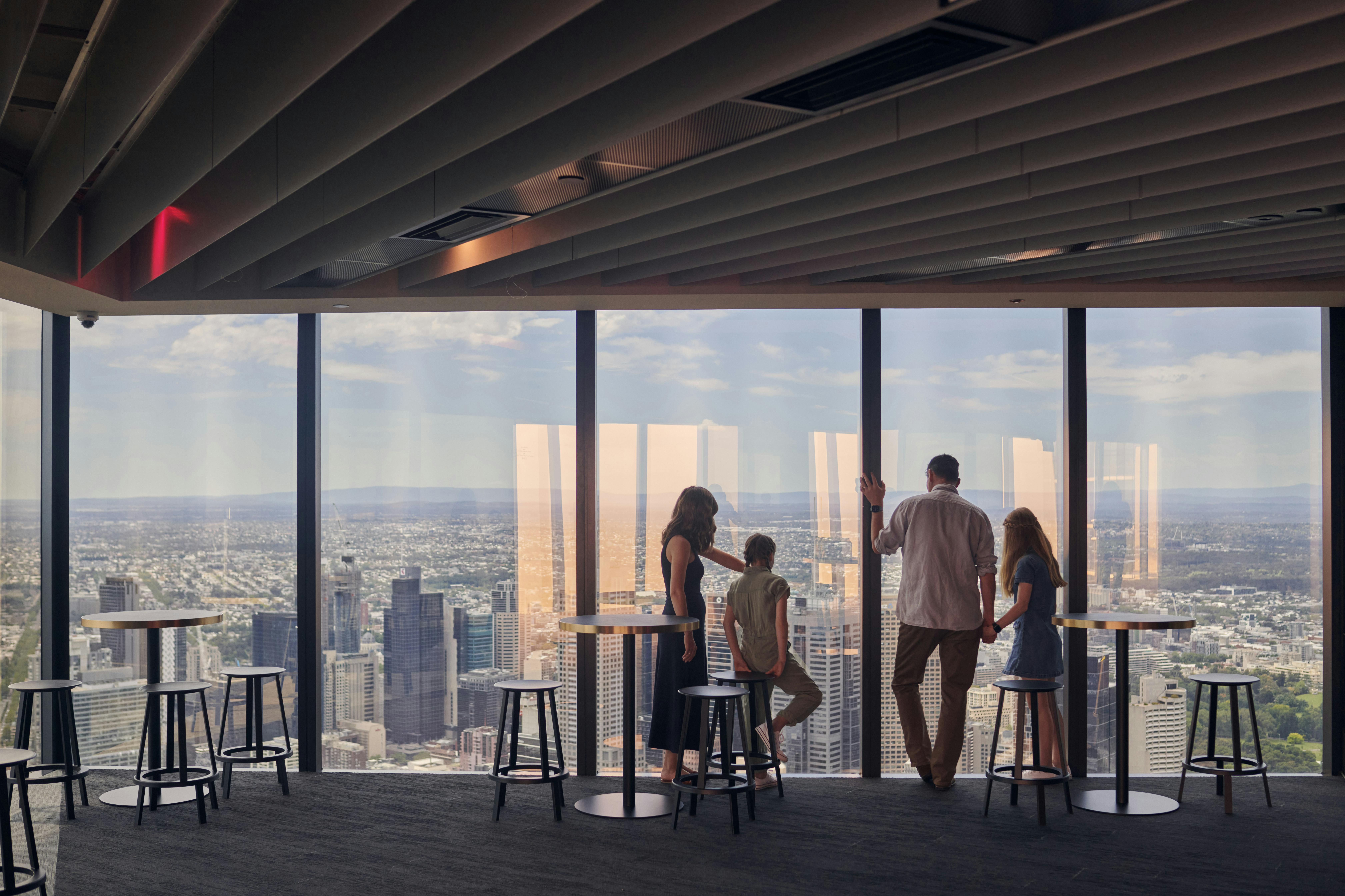 Four people stand by floor-to-ceiling windows in a high-rise building, overlooking a cityscape with tall buildings and scattered clouds.