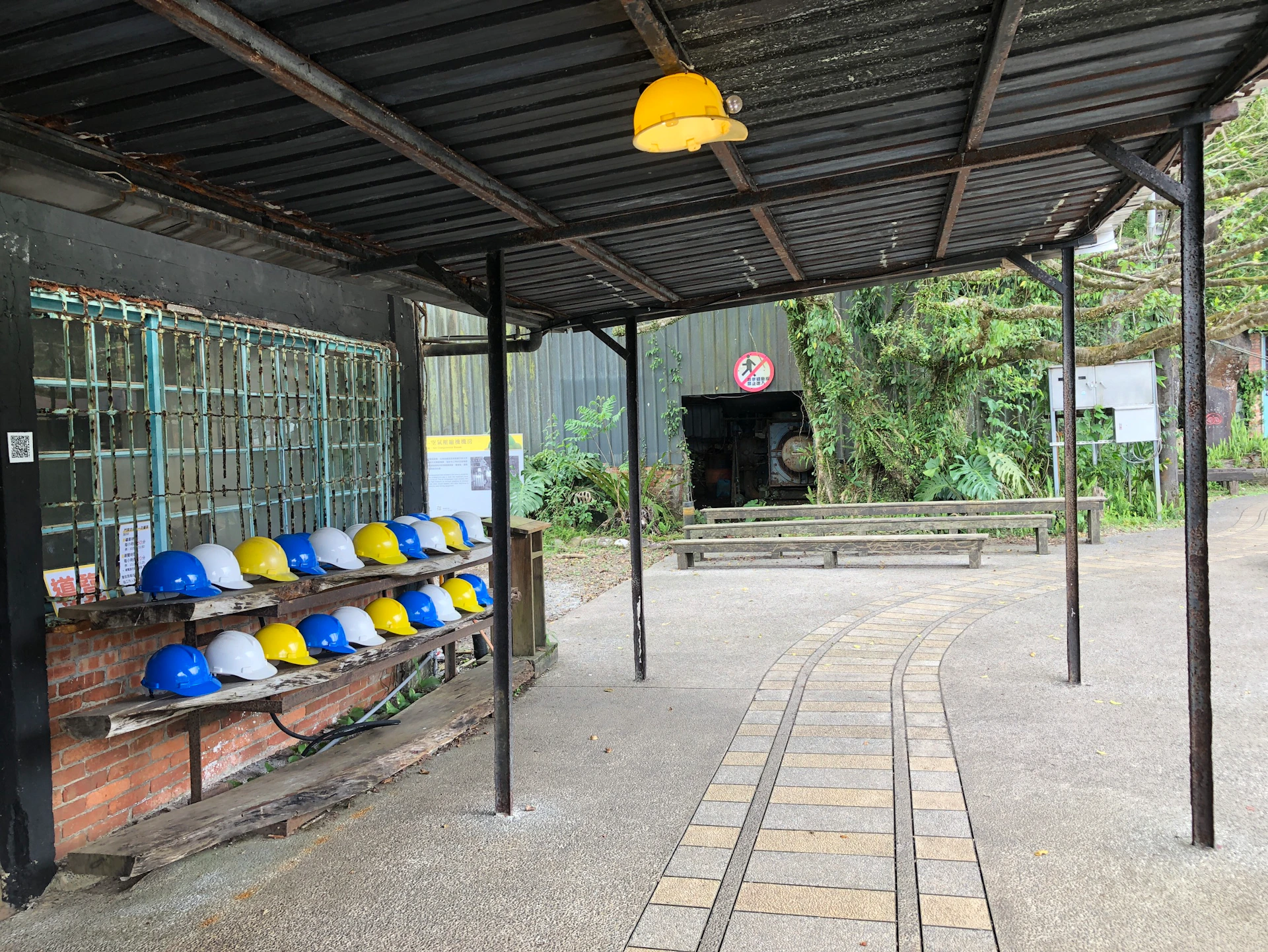 A covered area with a row of yellow and blue helmets on shelves, a pathway, and greenery in the background.