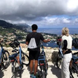 Fyra personer står med cyklar med utsikt över en naturskön kuststad under en molnig himmel.