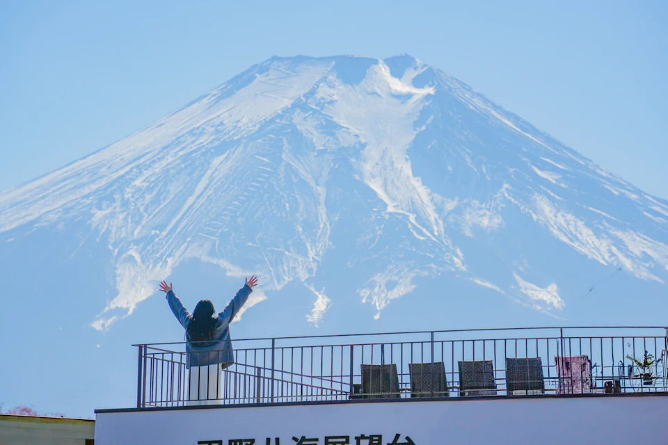 A person raising their arms stands on a balcony, with a clear view of a snow-capped mountain under a blue sky.