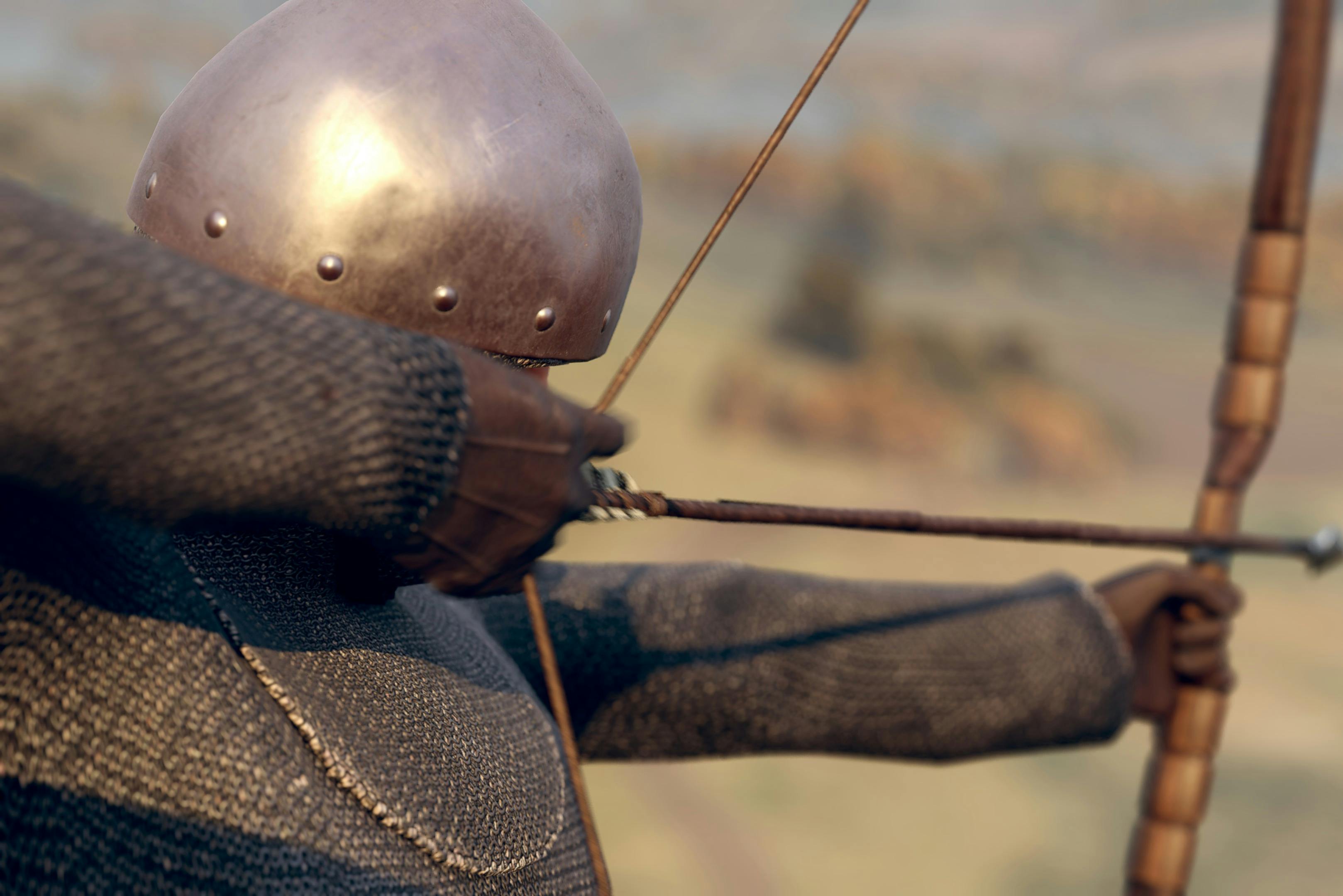 A person in chainmail and a metal helmet draws a bow, aiming into the distance with blurred landscape in the background.