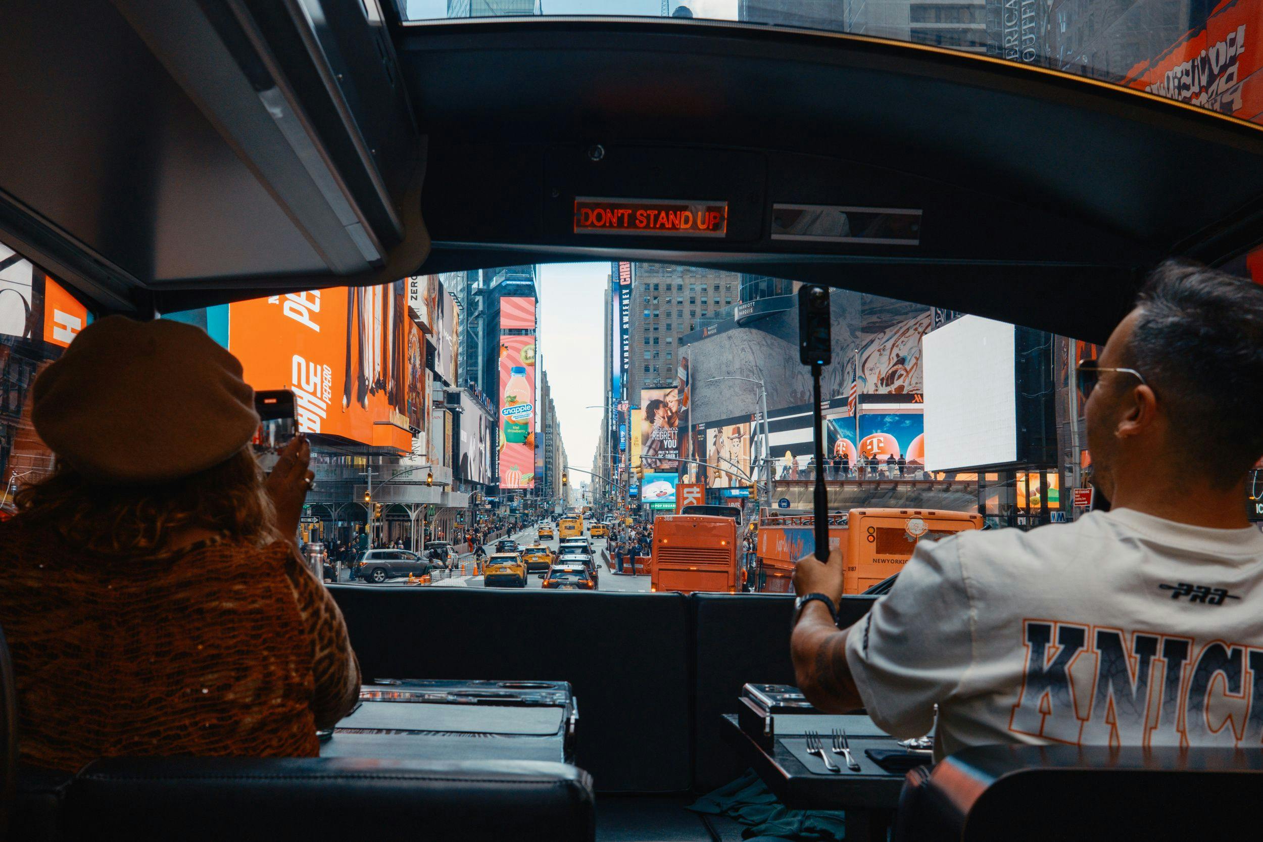 Vue de Times Square depuis l'intérieur d'un bus touristique ; panneaux publicitaires colorés, taxis jaunes et deux personnes au premier plan.