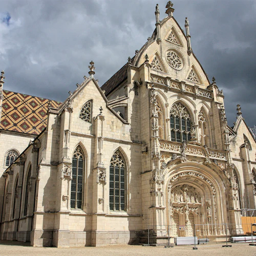Gotisk kyrka med invecklad fasad, välvda fönster och färgglatt mönstrat tak. Överskådlig himmel, få människor i förgrunden.
