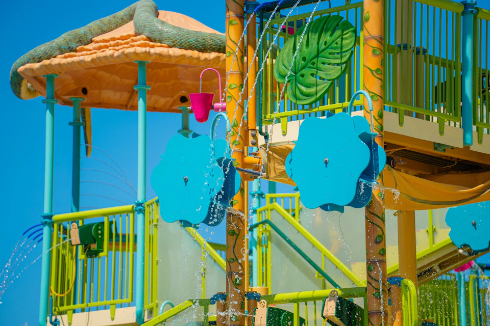 Colorful water playground with blue flower-shaped sprayers, yellow stairs, and green accents under a sunny sky.