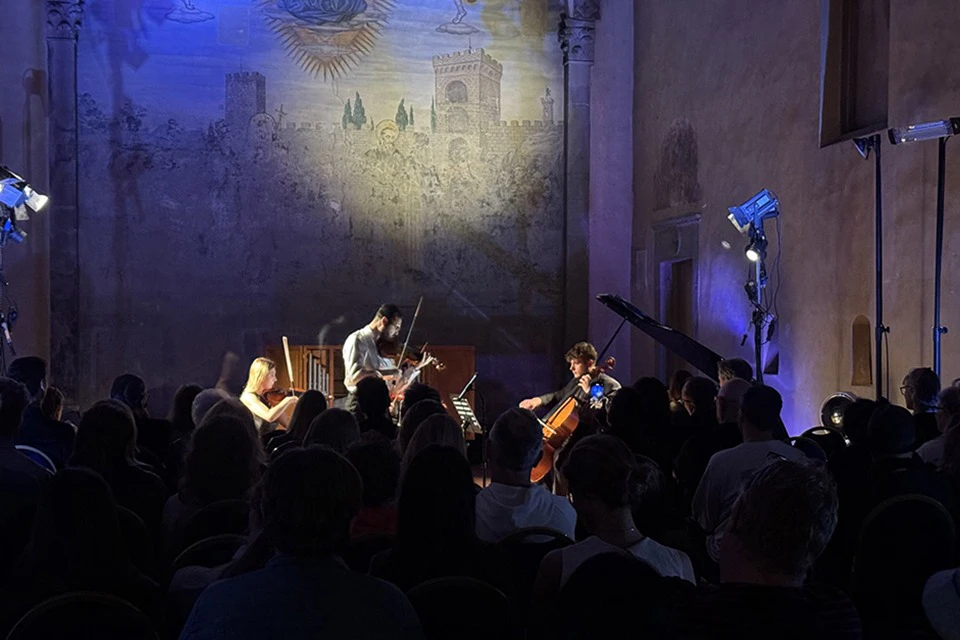 A small musical ensemble performs on stage in a dimly lit room with a painted wall and an audience in the foreground.