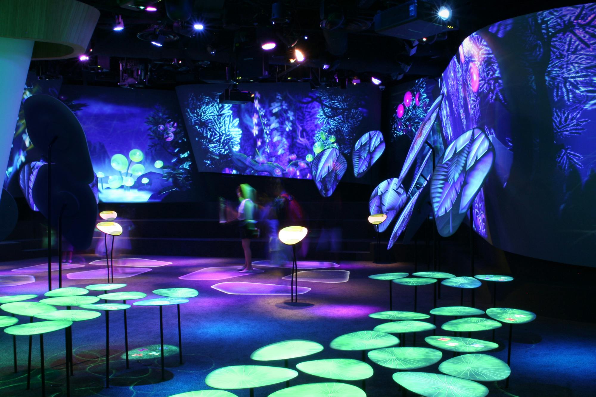 Tmavá místnost s neony osvětlenými projekcemi rostlin a hub s kruhovými světelnými instalacemi a abstraktními vzory. V pozadí jsou rozmazané postavy.