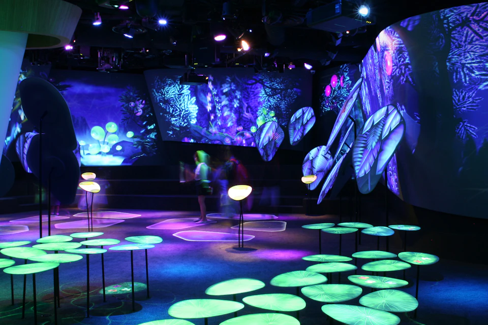 A dark room with neon-lit projections of plants and fungi, featuring circular light installations and abstract designs. There are blurred figures in the background.