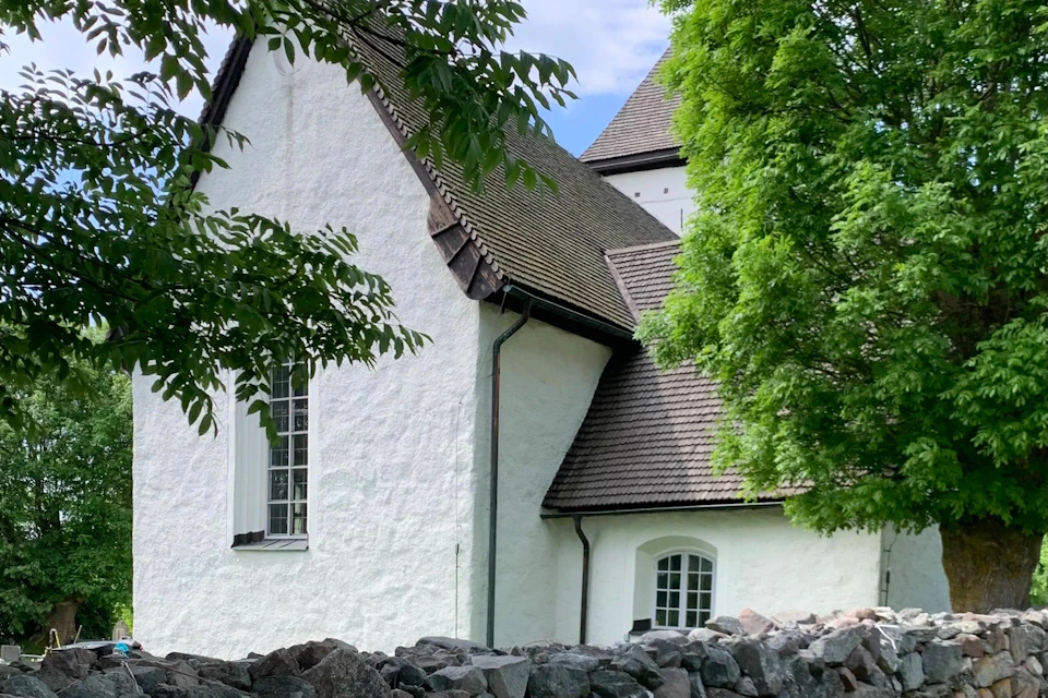 A white building with steep, brown shingled roofs, partially obscured by green trees and rocks in the foreground.