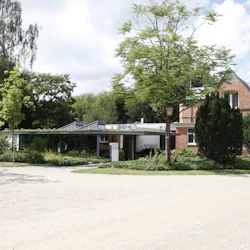 En modern glasbyggnad i anslutning till ett traditionellt tegelhus omgivet av frodig grönska och träd.