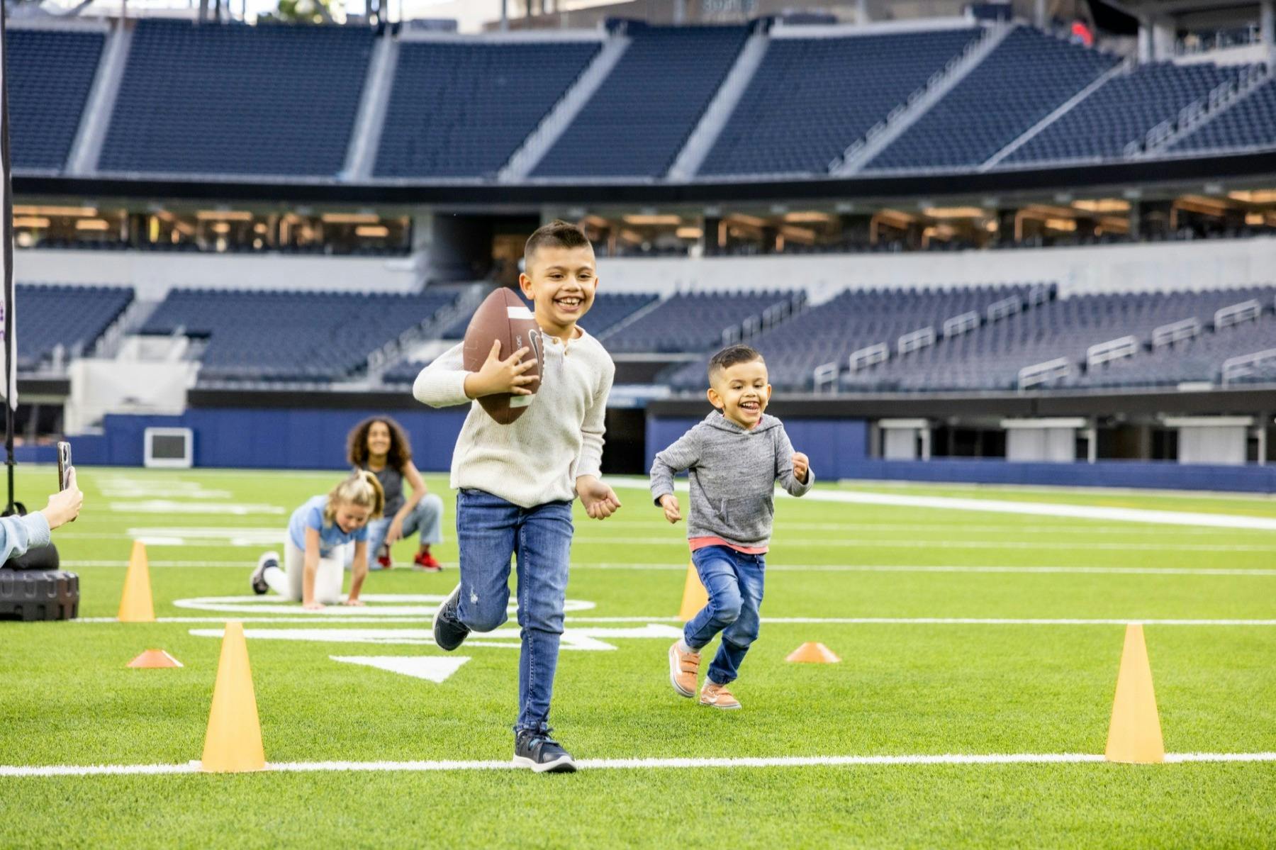 Children playing football on a stadium field, running near orange cones with a football in hand, and one crouching behind.