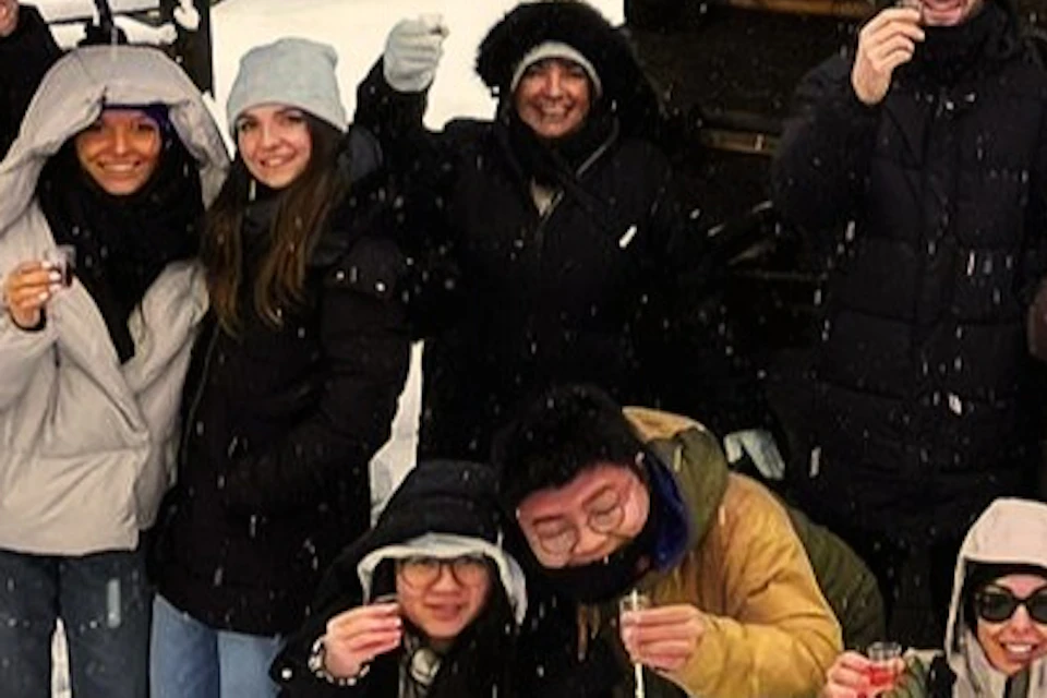 A group of people dressed in winter clothing posing outside a snow-covered wooden cabin in a snowy landscape.