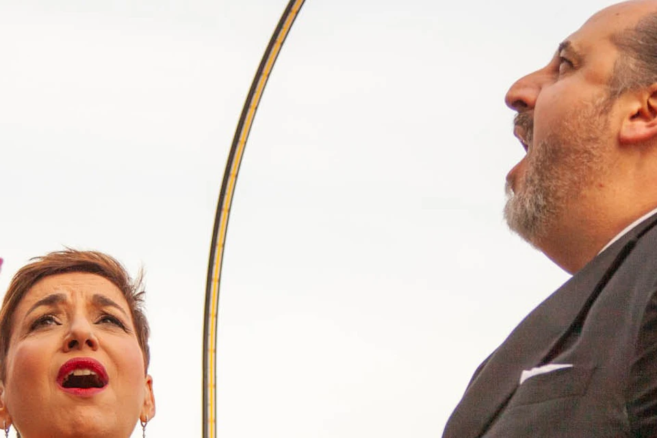 Two people performing passionately, one in a red dress and the other in a suit, against an outdoor backdrop.