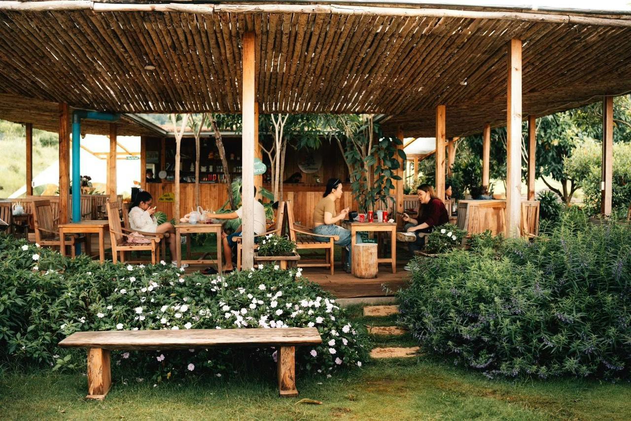 People sitting and talking at wooden tables in a garden cafe surrounded by plants and flowers under a bamboo roof.
