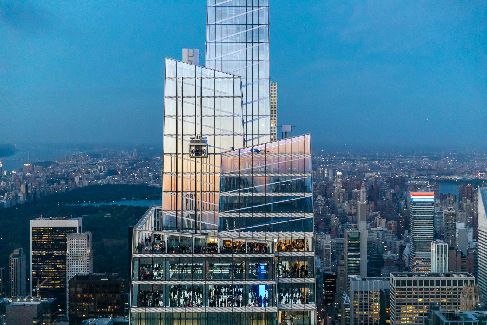 A modern skyscraper with reflective glass panels and an observation deck filled with people, set against a sprawling cityscape at dusk.