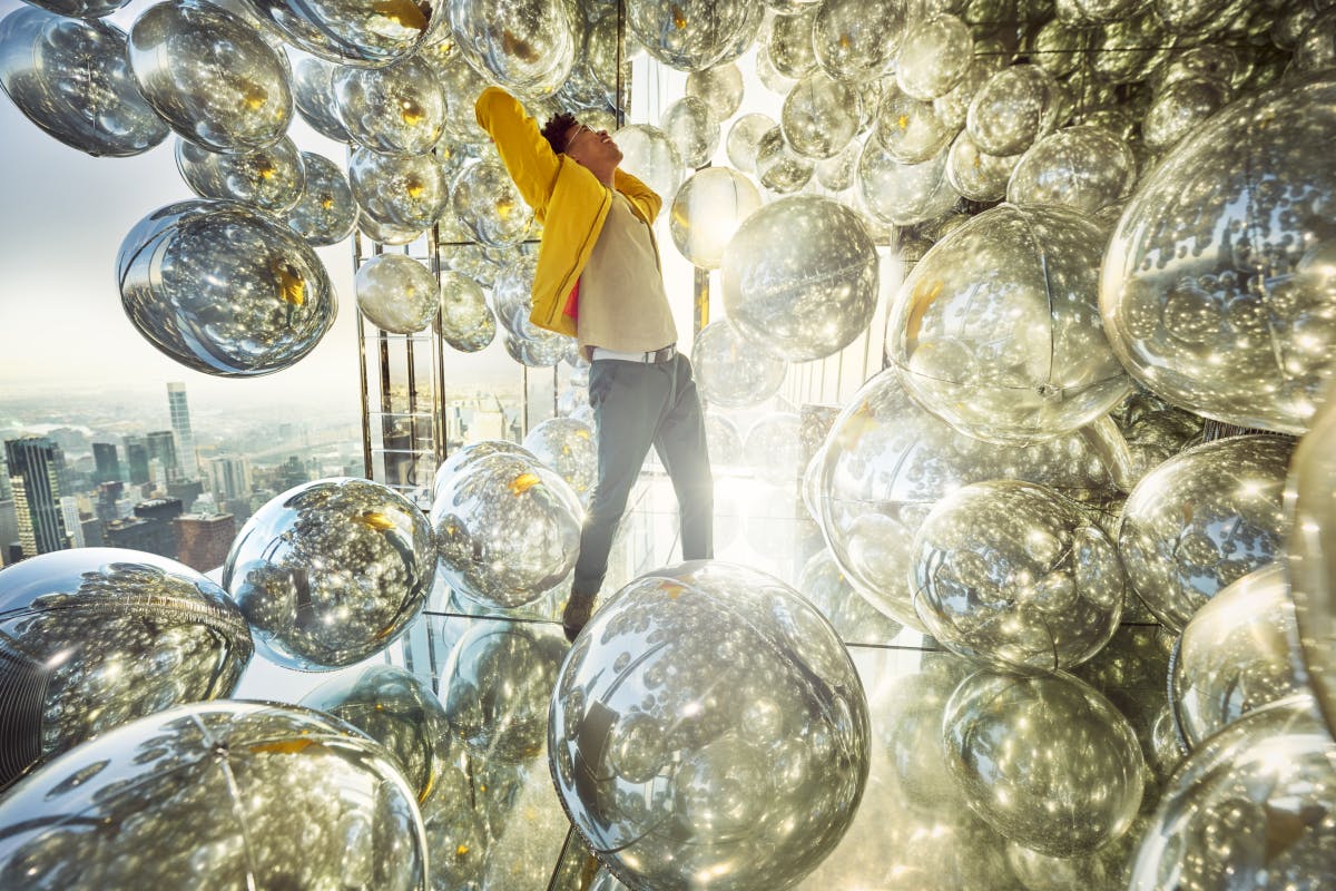 Ένα άτομο στέκεται ανάμεσα σε πολλές αντανακλαστικές ασημένιες σφαίρες, που εκτείνονται με τα χέρια υψωμένα, σε ένα καλά φωτισμένο δωμάτιο με ένα αστικό τοπίο ορατό έξω.