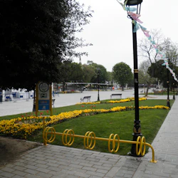En park med gula blommor, bänkar, en cykelhållare och lyktstolpar utsmyckade med flaggor. En person går på den asfalterade vägen.