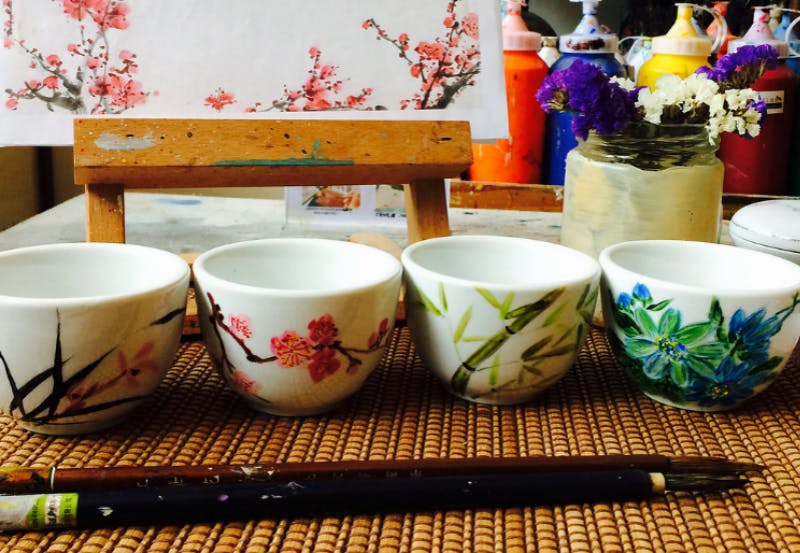 四个小陶瓷杯，编织垫上有花卉图案，前面有一把画笔，背景是一幅鲜花画。