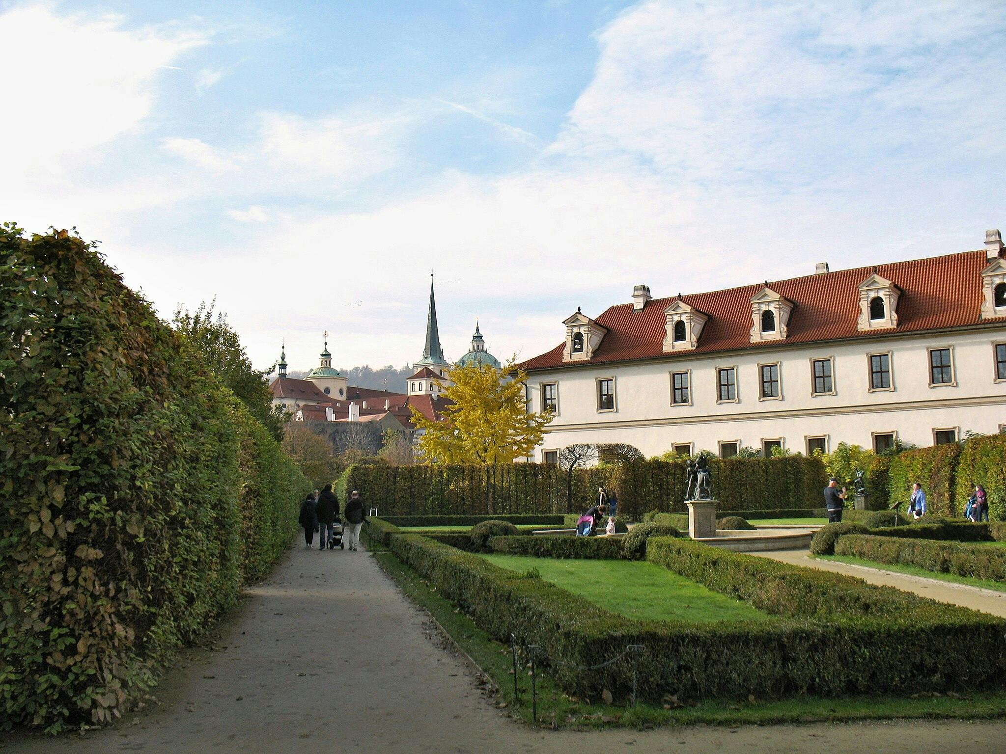 Manicured garden walkway with trimmed hedges and a historical building. People strolling and statues visible in the distance.