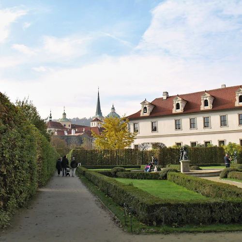 Välskött trädgårdsgång med klippta häckar och en historisk byggnad. Människor som promenerar och statyer som syns på avstånd.