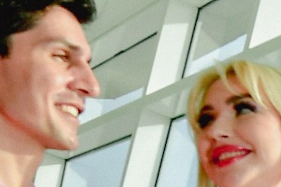 A couple stands in a modern glass-walled room with a scenic view, the woman pointing outside while smiling.