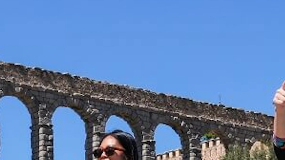 Four people stand in front of an ancient stone aqueduct under a clear blue sky with buildings and flowers in the background.