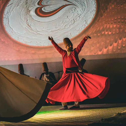 A person in a red flowing outfit performs a traditional spinning dance, with musicians and a projected design in the background.