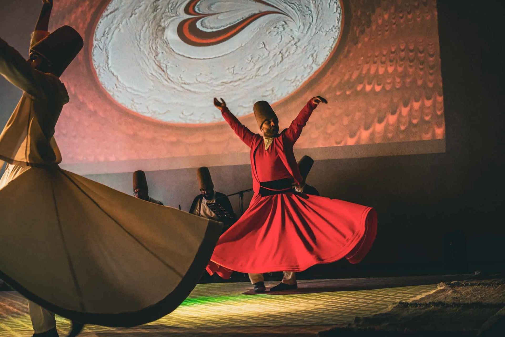 A person in a red flowing outfit performs a traditional spinning dance, with musicians and a projected design in the background.